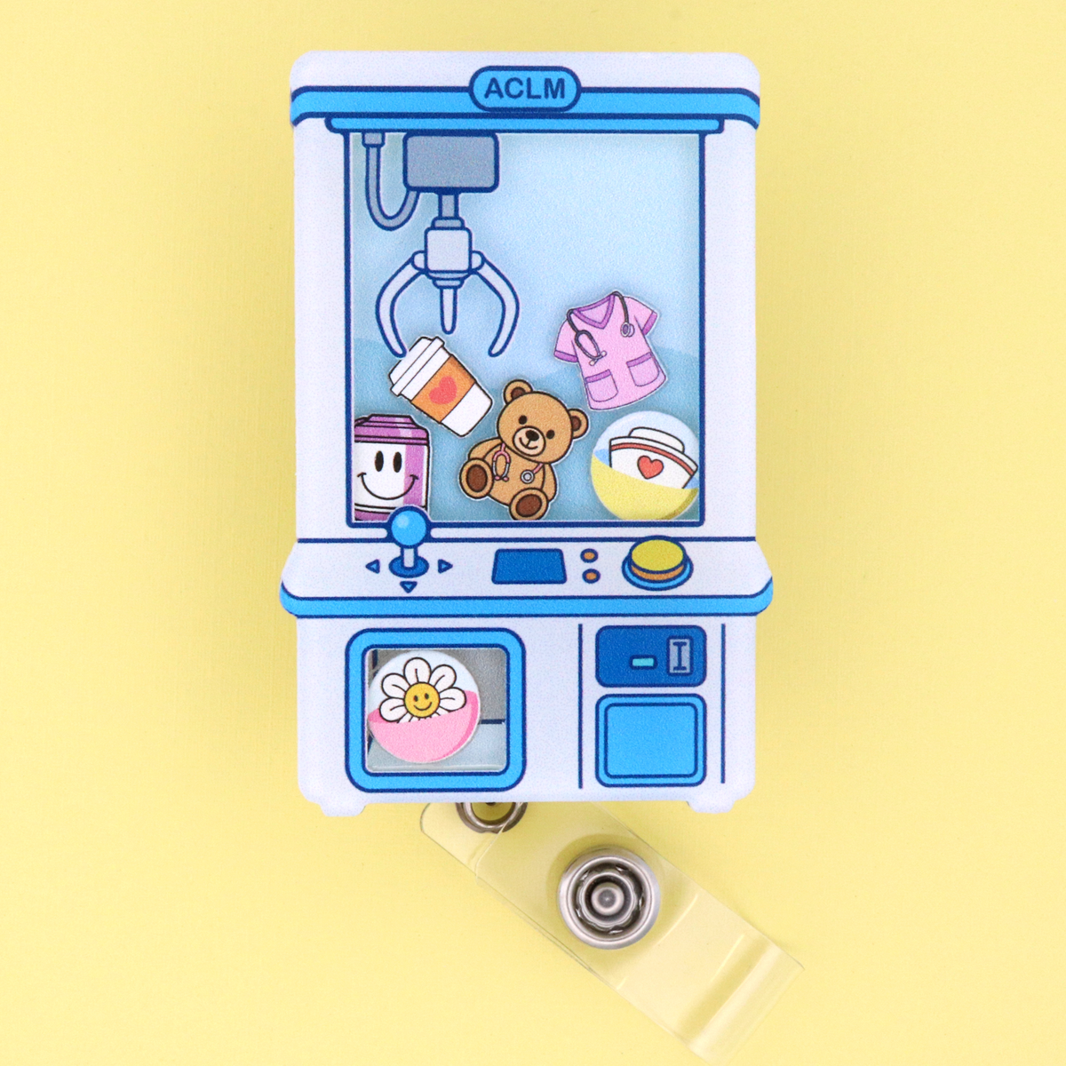 A miniature claw machine charm with a retractable badge reel, featuring cute pastel-colored toy items inside, set against a solid pale yellow background.